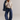 A woman modeling a cream V-neck knit sweater, blue wide-leg jeans, and olive-brown slip-on shoes, with a blue denim jacket draped over her shoulder, against a plain light gray studio background.