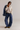 A woman modeling a cream V-neck knit sweater, blue wide-leg jeans, and olive-brown slip-on shoes, with a blue denim jacket draped over her shoulder, against a plain light gray studio background.