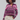 A woman with curly dark hair wearing a cropped, long-sleeve knit cardigan with horizontal stripes in purple, pink, and beige, paired with cream-colored pants, against a light gray studio background.