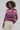 A woman with curly dark hair wearing a cropped, long-sleeve knit cardigan with horizontal stripes in purple, pink, and beige, paired with cream-colored pants, against a light gray studio background.