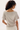 A woman with curly dark hair is shown from the back wearing a light beige short-sleeve top with a wide boat neckline and white bottoms, against a plain white studio background.