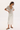 A woman modeling a light beige short-sleeve cowl-neck top and a white midi skirt with lace trim, paired with dark brown flip-flop sandals, against a plain white studio background.