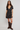 A woman modeling a short-sleeve black wrap-style mini dress with a beige and white floral print, paired with dark brown knee-high leather boots, against a plain light gray studio background.