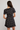 A woman shown from the back wearing a short-sleeve black dress with a beige and white floral pattern and a ruffled hem, set against a plain light gray background.