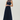 A woman modeling a sleeveless navy blue maxi dress with a sheer mesh high neckline and a pleated skirt, paired with black pointed flats, against a light gray studio background.
