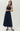 A woman modeling a sleeveless navy blue maxi dress with a sheer mesh high neckline and a pleated skirt, paired with black pointed flats, against a light gray studio background.