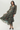 A woman modeling a long-sleeved midi dress with a green base and a red floral print, featuring a tie waist and a flowing ruffled hem, set against a plain light gray background.
