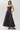 A woman models a strapless floor-length gown with a dark floral jacquard pattern in deep burgundy and black, featuring a defined waist and side pockets, set against a plain light gray studio background.