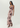 A woman modeling a sleeveless, floor-length pink maxi dress with a dark floral print, photographed against a plain light gray studio background.