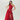 A woman modeling a floor-length red one-shoulder evening gown with a ruched bodice, draped skirt detail, and high front slit, photographed against a clean light gray studio background.