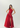 A woman modeling a floor-length red one-shoulder evening gown with a ruched bodice, draped skirt detail, and high front slit, photographed against a clean light gray studio background.