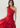 A woman modeling a bright red one-shoulder draped gown against a plain light gray studio background.