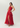 A woman modeling a floor-length red one-shoulder gown with a draped bodice, high slit, and flowing skirt against a plain light gray studio background.
