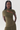 A model with long black braids wears a textured olive green wrap-style dress with a draped shoulder and a fitted bodice, photographed against a plain light gray studio background.