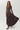A woman modeling a long-sleeve brown maxi dress with a sheer mesh neckline and a flowing satin skirt, posed against a light gray studio background.
