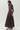 A woman modeling a long-sleeve brown maxi dress with a pleated skirt, shown from a side-back angle against a plain light gray studio background.