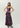 A woman modeling a strapless floor-length gown with a dark base and large pink, purple, and olive floral print, posed against a light gray studio background.