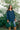 A woman stands outdoors in front of leafy green trees, wearing a navy blue plaid coat with brown collar and cuff trim, blue jeans, tan boots, and holding a dark green handbag.