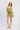 A woman modeling a two-piece olive green romper set with a ruffled smocked top and matching shorts, paired with beige woven flat shoes, against a light gray studio background.
