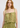 A woman wearing a light olive green ruffled camisole top with a smocked waist, shown against a plain light gray studio background.