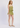 A woman modeling a light olive green two-piece set consisting of a ruffled spaghetti-strap top and matching smocked ruffle shorts, paired with beige woven flat shoes, against a plain light gray studio background.