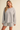 A woman modeling a light heather gray knit lounge set consisting of a crewneck sweatshirt and matching shorts, photographed against a solid beige studio background.