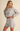 A woman modeling a light heather gray knit lounge set consisting of a cropped long-sleeve crewneck sweater and matching high-waisted shorts, set against a solid warm beige background.