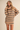A woman wearing an oversized beige knit sweater dress with horizontal navy stripes, posed against a solid beige studio background.