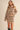 A woman wearing an oversized beige knit sweater dress with horizontal navy blue stripes, styled with black ankle boots against a solid beige studio background.