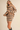 A woman models an oversized beige knit sweater dress with horizontal black stripes, styled with black ankle boots against a solid beige background.