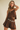 A woman modeling a brown faux-suede two-piece outfit consisting of a sleeveless ruffled crop top and matching pleated shorts, styled with a silver chain belt featuring oval medallion charms, against a plain beige background.
