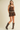 A woman modeling a brown suede-like fringed romper with a silver concho belt, paired with brown leather cowboy boots, against a solid beige studio background.