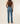 Rear view of a model wearing medium-wash blue straight-leg jeans, a brown ribbed tank top, and gray slip-on shoes against a warm off-white studio background.