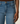 Close-up of a person wearing medium-wash blue denim jeans, showing the front waistband, belt loops, button, pocket, and hand resting beside the leg against a plain light background.