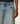 Close-up of a person wearing light blue denim jeans with a faded wash, showing the front pocket, waistband, and hand resting near the thigh against a plain light background.