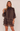 A woman modeling a dark brown tiered ruffle mini dress with a high mock neck and sheer long sleeves, set against a plain light beige background.