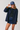 A woman modeling a navy blue button-front overshirt with brown collar and cuff trim, worn over a black top and denim skirt, while wearing a navy baseball cap with white text.