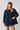 A woman modeling a navy blue button-up jacket with brown corduroy collar and cuff trim, worn over a black top and a denim mini skirt, with a matching blue cap.