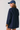 A woman shown from the back wearing an oversized navy blue shirt jacket with brown suede-like cuffs and collar trim, paired with a navy baseball cap, against a light gray studio background.
