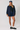 A woman modeling a navy blue utility jacket with brown corduroy collar and cuffs, paired with a black top, denim skirt, navy baseball cap, white socks, and black loafers, against a light gray studio background.