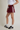 A model wearing a pair of burgundy athletic shorts with white side stripes, paired with a white crop top, white socks, and black Adidas sneakers, shown from the waist down against a neutral studio background.