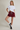 A model posing in a studio setting wearing a white cropped t-shirt, a burgundy drawstring mini skirt, white socks, and black Adidas sneakers against a light beige background.