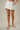 A model wearing a white denim mini skirt with pleated details and front pockets, paired with clear-strap heeled sandals, against a neutral beige background.