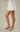 A model wearing a pair of white high-waisted shorts with pleated detailing and a white top, paired with nude heeled mule sandals, against a solid beige studio background.