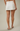 A model from the back wearing a white high-waisted mini skirt with pleated details and side pockets, paired with beige heeled sandals against a solid beige studio background.