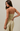 A woman modeling a strapless bandeau-style jumpsuit with horizontal stripes in olive green and cream, shown from a three-quarters back view against a solid light beige background.