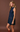 A woman modeling a sleeveless dark blue denim dress with front pockets, standing against a solid brown studio background.