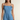 A woman modeling a light blue denim romper with thin shoulder straps, tie details at the shoulders, a square neckline, and a ruffled hem against a plain beige background.