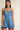 A woman modeling a light blue denim romper with thin shoulder straps, tie details at the shoulders, a square neckline, and a ruffled hem against a plain beige background.
