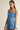 A woman modeling a light blue denim camisole dress with tie straps and a ruffled hem against a plain beige studio background.